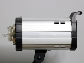 A silver studio light with black accents mounted on a support bracket. The cylindrical shape features small vents and a clear bulb extending from the left side.