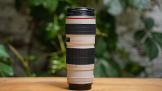 A large, professional camera lens with black rubber grips and white accents, placed vertically on a wooden surface. The background features green foliage, adding a natural and relaxed atmosphere.