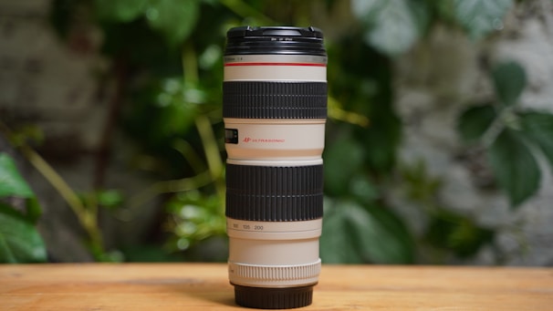 A large, professional camera lens with black rubber grips and white accents, placed vertically on a wooden surface. The background features green foliage, adding a natural and relaxed atmosphere.