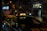 A taxi with an illuminated sign reading 'Taxi Parisien' is parked on a street at night. The vehicle has a glossy appearance, possibly due to rain. Surrounding the taxi, city lights and reflections are visible from nearby buildings and a bus on the move.