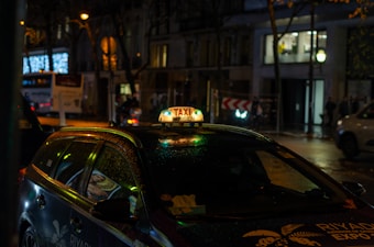 A taxi with an illuminated sign reading 'Taxi Parisien' is parked on a street at night. The vehicle has a glossy appearance, possibly due to rain. Surrounding the taxi, city lights and reflections are visible from nearby buildings and a bus on the move.