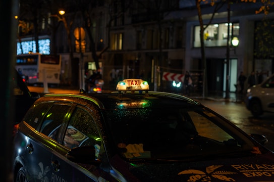 A taxi with an illuminated sign reading 'Taxi Parisien' is parked on a street at night. The vehicle has a glossy appearance, possibly due to rain. Surrounding the taxi, city lights and reflections are visible from nearby buildings and a bus on the move.