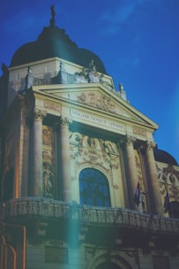 A grand, ornately decorated building facade features intricate carvings and columns. It has an inscription that reads 'Magyar Nemzeti Színház.' The architectural style includes decorative statues and embellishments under a blue sky.