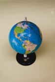 A small globe displaying the geographical layout of North and South America against a solid light yellow background. The globe is mainly blue with green, yellow, and brown landmasses and is mounted on a black base.
