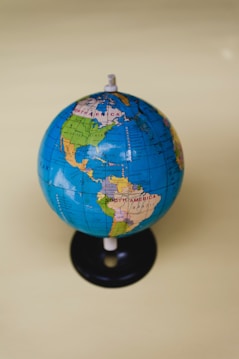 A small globe displaying the geographical layout of North and South America against a solid light yellow background. The globe is mainly blue with green, yellow, and brown landmasses and is mounted on a black base.