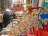 A vibrant market stall displays a variety of colorful items including handcrafted camels, souvenirs, clothing, musical instruments, and ornate textiles. Shoppers gather around, browsing and interacting with the vendor, creating a lively atmosphere.