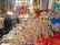 A vibrant market stall displays a variety of colorful items including handcrafted camels, souvenirs, clothing, musical instruments, and ornate textiles. Shoppers gather around, browsing and interacting with the vendor, creating a lively atmosphere.