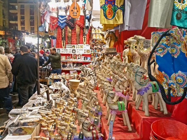 A vibrant market stall displays a variety of colorful items including handcrafted camels, souvenirs, clothing, musical instruments, and ornate textiles. Shoppers gather around, browsing and interacting with the vendor, creating a lively atmosphere.