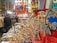 A vibrant market stall displays a variety of colorful items including handcrafted camels, souvenirs, clothing, musical instruments, and ornate textiles. Shoppers gather around, browsing and interacting with the vendor, creating a lively atmosphere.