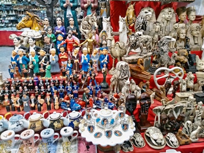 A vibrant marketplace stall displaying a diverse collection of small statues, figurines, and decorative items. Prominent among the items are figures resembling famous leaders, ancient Egyptian statues, animals, and various ceramic mugs and teapots with portraits. The display is colorful and densely packed, with items placed on red fabric-covered shelves.