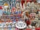 A vibrant marketplace stall displaying a diverse collection of small statues, figurines, and decorative items. Prominent among the items are figures resembling famous leaders, ancient Egyptian statues, animals, and various ceramic mugs and teapots with portraits. The display is colorful and densely packed, with items placed on red fabric-covered shelves.