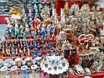 A vibrant marketplace stall displaying a diverse collection of small statues, figurines, and decorative items. Prominent among the items are figures resembling famous leaders, ancient Egyptian statues, animals, and various ceramic mugs and teapots with portraits. The display is colorful and densely packed, with items placed on red fabric-covered shelves.