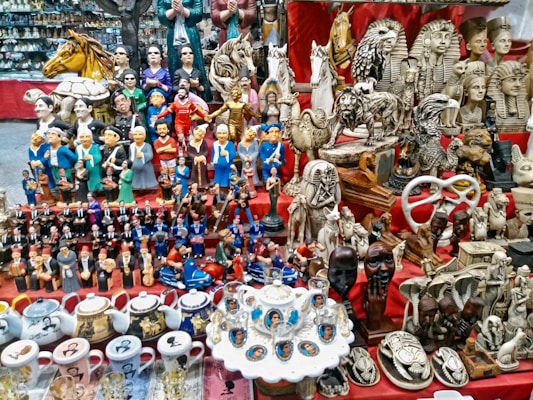 A vibrant marketplace stall displaying a diverse collection of small statues, figurines, and decorative items. Prominent among the items are figures resembling famous leaders, ancient Egyptian statues, animals, and various ceramic mugs and teapots with portraits. The display is colorful and densely packed, with items placed on red fabric-covered shelves.