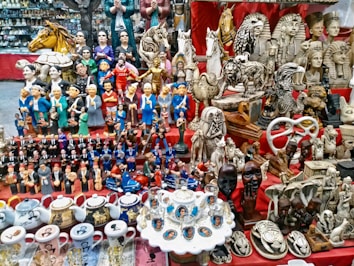 A vibrant marketplace stall displaying a diverse collection of small statues, figurines, and decorative items. Prominent among the items are figures resembling famous leaders, ancient Egyptian statues, animals, and various ceramic mugs and teapots with portraits. The display is colorful and densely packed, with items placed on red fabric-covered shelves.