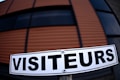 A sign with the word 'VISITEURS' is prominently displayed in clear, bold black letters against a white background. The setting includes a large structure in the background with horizontal stripes in shades of brown and black, suggesting a modern architectural design.