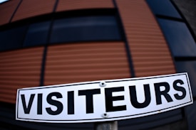 A sign with the word 'VISITEURS' is prominently displayed in clear, bold black letters against a white background. The setting includes a large structure in the background with horizontal stripes in shades of brown and black, suggesting a modern architectural design.
