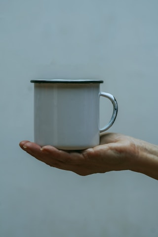 An enamel mug with a dark rim is held by a hand against a muted, neutral background.