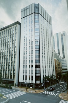 A modern urban scene with tall, sleek buildings. The architecture is characterized by a glass facade and sharp lines, with a prominent intersection in the foreground, marked by clear crosswalks. Trees and greenery are scattered around the street, adding a touch of nature to the cityscape.