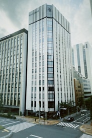 A modern urban scene with tall, sleek buildings. The architecture is characterized by a glass facade and sharp lines, with a prominent intersection in the foreground, marked by clear crosswalks. Trees and greenery are scattered around the street, adding a touch of nature to the cityscape.