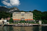 A grand hotel building with a classic architectural design is situated by the water, surrounded by lush greenery and small mountains in the background. The structure features multiple floors with balconies and an elegant facade. There is a dock and sun loungers with umbrellas in front of the hotel along the water's edge.
