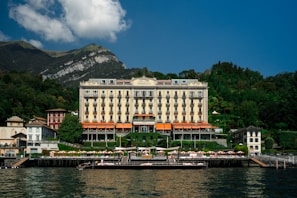 A grand hotel building with a classic architectural design is situated by the water, surrounded by lush greenery and small mountains in the background. The structure features multiple floors with balconies and an elegant facade. There is a dock and sun loungers with umbrellas in front of the hotel along the water's edge.