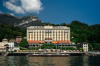 A grand hotel building with a classic architectural design is situated by the water, surrounded by lush greenery and small mountains in the background. The structure features multiple floors with balconies and an elegant facade. There is a dock and sun loungers with umbrellas in front of the hotel along the water's edge.