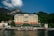 A grand hotel building with a classic architectural design is situated by the water, surrounded by lush greenery and small mountains in the background. The structure features multiple floors with balconies and an elegant facade. There is a dock and sun loungers with umbrellas in front of the hotel along the water's edge.