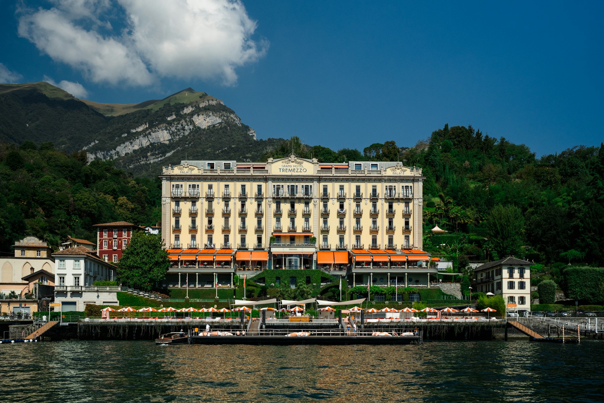 A grand hotel building with a classic architectural design is situated by the water, surrounded by lush greenery and small mountains in the background. The structure features multiple floors with balconies and an elegant facade. There is a dock and sun loungers with umbrellas in front of the hotel along the water's edge.