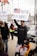 A peaceful protest is taking place with a man holding a sign featuring an American flag stating 'Not in Our Names'. A young child sits in a stroller holding two American flags. There are other people in the background holding signs, indicating a group demonstration. The scene is set outdoors on a city street with parked cars and palm trees visible.