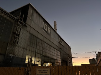 An industrial building with corrugated metal walls and large windows. A ladder is attached to the side of the building, and a smokestack protrudes from the roof. There is a barbed wire fence in the foreground with a 'private property, no trespassing' sign affixed to it. The sky in the background displays a gradient from dark blue to orange near the horizon, indicating either sunrise or sunset.