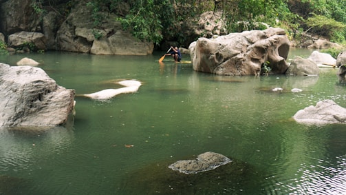 A serene natural pond surrounded by large boulders and lush greenery. A person is seen in the distance engaging with the water, possibly fishing or maneuvering a wooden stick. The water is calm and reflects the surrounding rocks and trees.