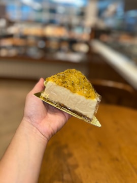 A hand is holding a slice of cheesecake topped with a glossy yellow fruit compote that appears to have seeds. The background shows a blurred image of what seems to be a bakery or café with various baked goods displayed.