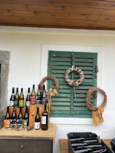 A rustic setting is arranged with several wine bottles displayed on a wooden surface. Above, there are two wreaths on a green shuttered window, one made of wine corks and the other of twigs. The backdrop is a white wall with a wooden ceiling.