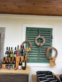 A rustic setting is arranged with several wine bottles displayed on a wooden surface. Above, there are two wreaths on a green shuttered window, one made of wine corks and the other of twigs. The backdrop is a white wall with a wooden ceiling.