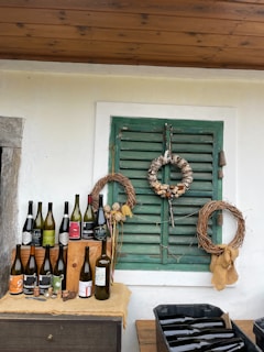 A rustic setting is arranged with several wine bottles displayed on a wooden surface. Above, there are two wreaths on a green shuttered window, one made of wine corks and the other of twigs. The backdrop is a white wall with a wooden ceiling.