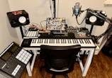A music production setup featuring a keyboard controller on a white desk, with a laptop covered in stickers. Surrounding the keyboard are studio monitors, a microphone on a stand, and various music production equipment like a control pad and mixing console. Cables are neatly organized, and there is a small red car model on top of one of the speakers.