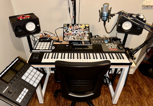 A music production setup featuring a keyboard controller on a white desk, with a laptop covered in stickers. Surrounding the keyboard are studio monitors, a microphone on a stand, and various music production equipment like a control pad and mixing console. Cables are neatly organized, and there is a small red car model on top of one of the speakers.