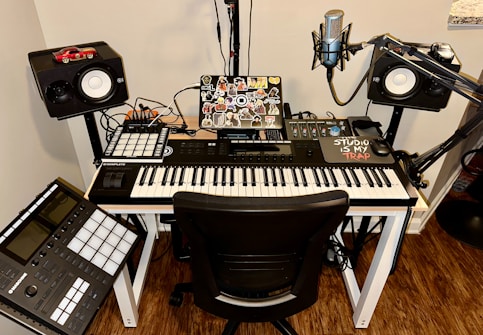 A music production setup featuring a keyboard controller on a white desk, with a laptop covered in stickers. Surrounding the keyboard are studio monitors, a microphone on a stand, and various music production equipment like a control pad and mixing console. Cables are neatly organized, and there is a small red car model on top of one of the speakers.