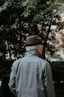 An elderly person stands outdoors, wearing a brown hat and a grey coat, with their back turned. They are surrounded by lush, green foliage and appear to be in a peaceful, natural setting.