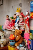 A collection of toys and figurines is arranged on a shelf. There are several dolls dressed in colorful outfits, a teddy bear, a superhero figure in a red and silver suit, and a small stuffed animal resembling a llama. Additional items include a ceramic figure and decorative jars. The background features a painted mural with bright colors.