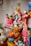 A collection of toys and figurines is arranged on a shelf. There are several dolls dressed in colorful outfits, a teddy bear, a superhero figure in a red and silver suit, and a small stuffed animal resembling a llama. Additional items include a ceramic figure and decorative jars. The background features a painted mural with bright colors.