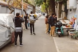 A narrow urban street lined with gray walls and overhanging trees. Several people are engaged in photography, capturing images of the area. A motorcycle is parked beside a small altar or display with various objects, including small statues. The scene feels bustling yet intimate.