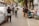 A narrow urban street lined with gray walls and overhanging trees. Several people are engaged in photography, capturing images of the area. A motorcycle is parked beside a small altar or display with various objects, including small statues. The scene feels bustling yet intimate.