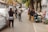 A narrow urban street lined with gray walls and overhanging trees. Several people are engaged in photography, capturing images of the area. A motorcycle is parked beside a small altar or display with various objects, including small statues. The scene feels bustling yet intimate.
