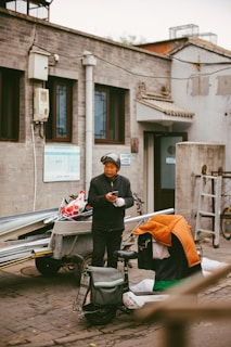 A person wearing a helmet and dark clothing stands beside a scooter, holding a phone. The scooter has a colorful cover draped over it and is next to a trailer filled with metal rods and a bag with a red and white pattern. The setting is an urban alley with grey brick buildings featuring traditional architectural elements like small roof tiles and decorative window frames.