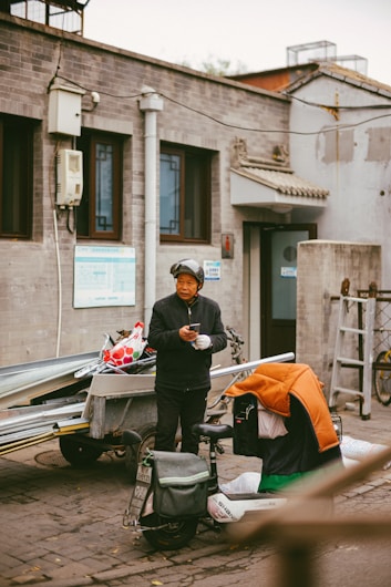 A person wearing a helmet and dark clothing stands beside a scooter, holding a phone. The scooter has a colorful cover draped over it and is next to a trailer filled with metal rods and a bag with a red and white pattern. The setting is an urban alley with grey brick buildings featuring traditional architectural elements like small roof tiles and decorative window frames.