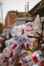 A cluttered pile of empty plastic bottles, primarily with red labels, appears stacked or heaped together outdoors against an urban backdrop.