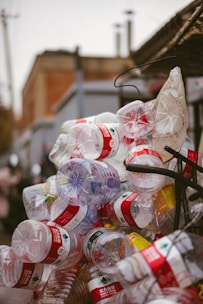 A cluttered pile of empty plastic bottles, primarily with red labels, appears stacked or heaped together outdoors against an urban backdrop.