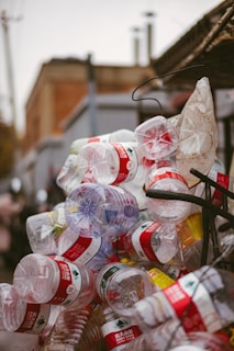 A cluttered pile of empty plastic bottles, primarily with red labels, appears stacked or heaped together outdoors against an urban backdrop.