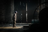 A solitary figure stands in a dimly lit industrial space, gazing towards a crack in a large, rusty door. The light seeping through creates a stark contrast against the dark interior, illuminating the person and casting long shadows. The scene presents an atmosphere of mystery and introspection.